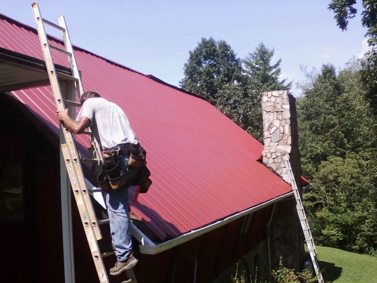 Roofer on a ladder beside a red metal roof and stone chimney — Soffit & Fascia Repair by Roofing Brothers in Jennings